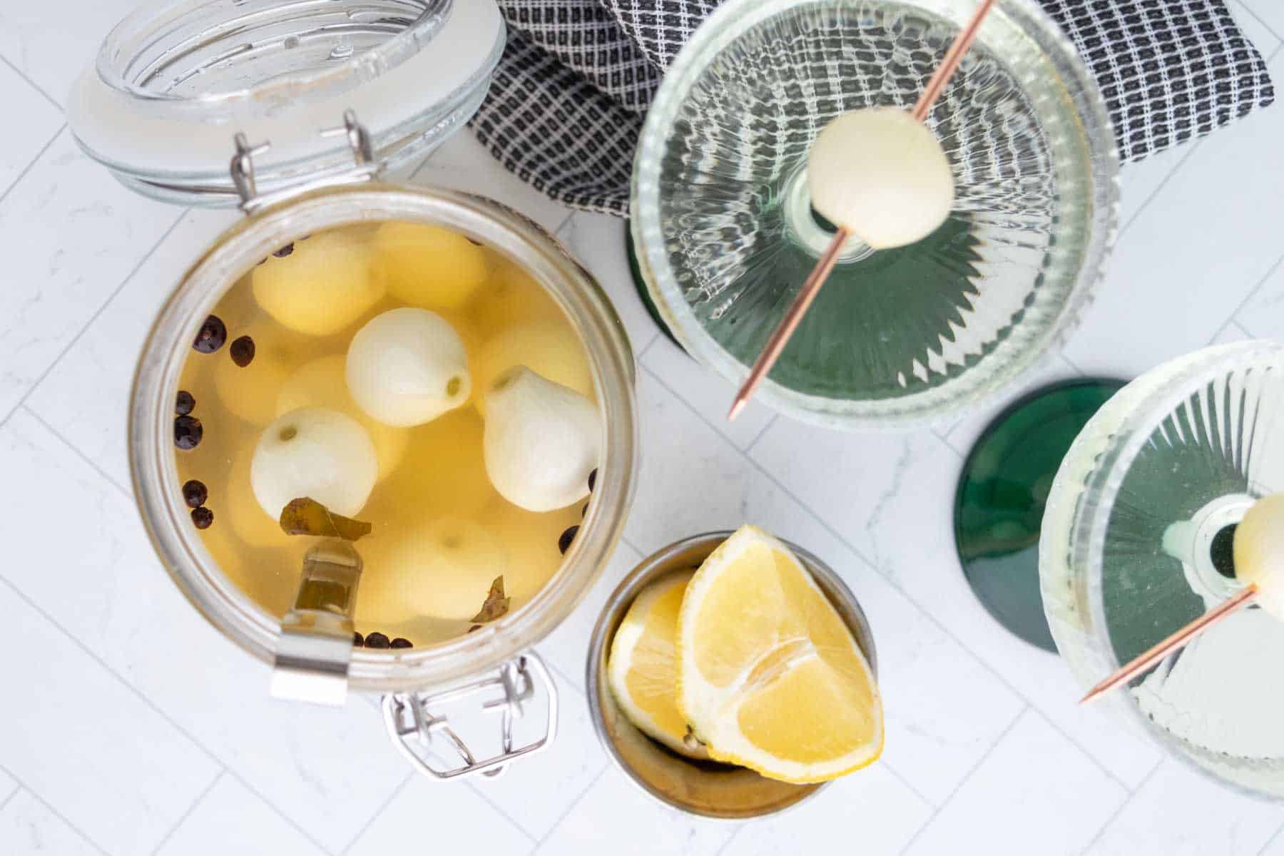 A jar of pickled onions with spices, two martini glasses each garnished with an onion, and a glass with lemon wedges on a white tiled surface.