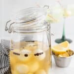 A glass jar filled with homemade cocktail onions in brine, with peppercorns, sits on a counter beside lemon wedges and two cocktail glasses.