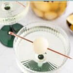 A cocktail glass with a single cocktail onion on a cocktail pick resting on the rim, with another similar glass and a jar of cocktail onions in the background.