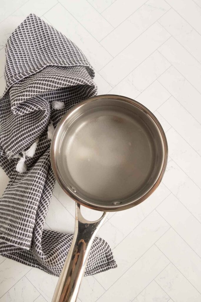 A stainless steel saucepan with water inside is placed on a white tiled surface next to a black and white checkered kitchen towel.