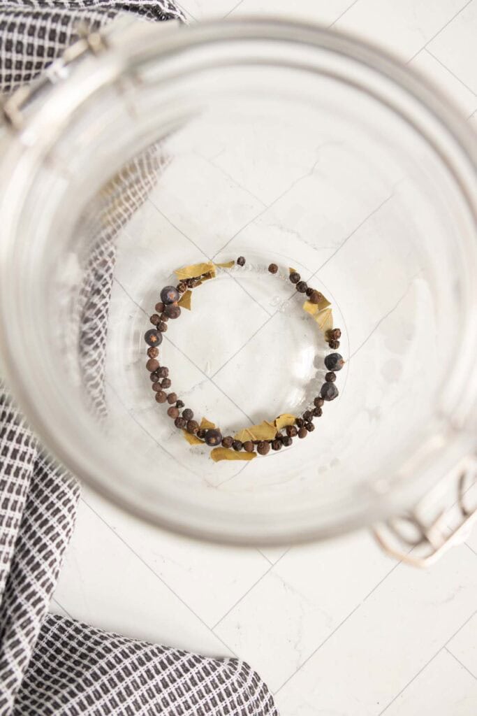 A glass jar containing a small amount of whole spices, including peppercorns and bay leaves, on a white tiled surface.