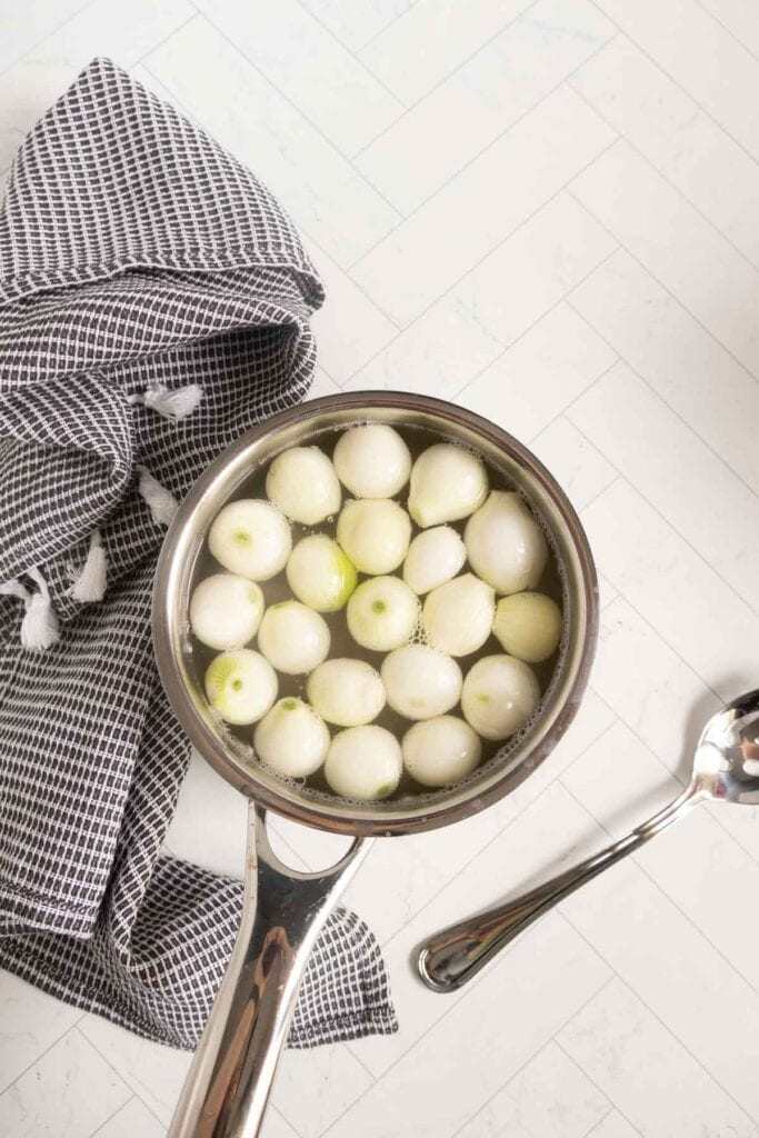 A saucepan filled with peeled pearl onions in water sits on a white countertop next to a checkered kitchen towel and a metal spoon.
