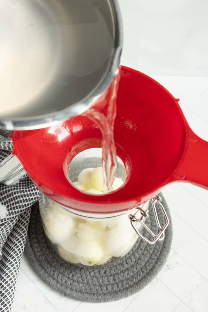 A pot pours liquid through a red funnel into a glass jar filled with peeled onions, resting on a gray mat next to a checkered towel.