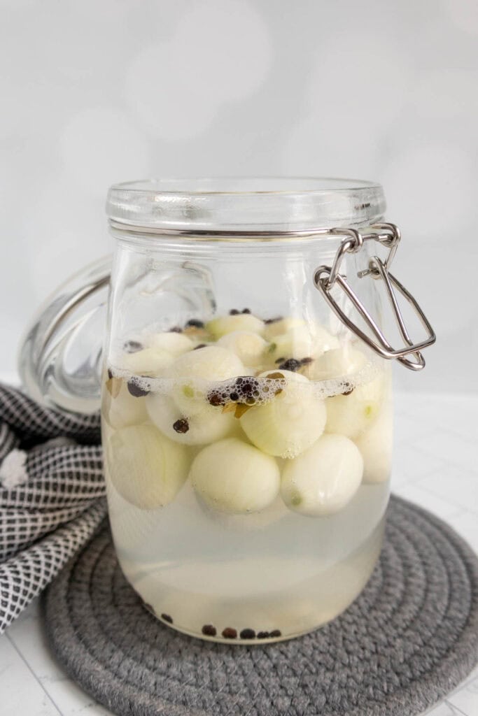 A glass jar filled with pickled pearl onions and peppercorns in brine, placed on a round gray mat with a checkered cloth nearby.