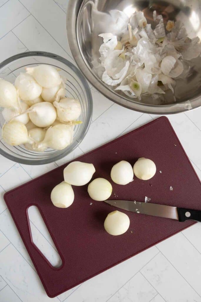 Peeled pearl onions on a cutting board with a knife, a bowl of peeled onions, and a metal bowl containing onion skins on a white countertop.