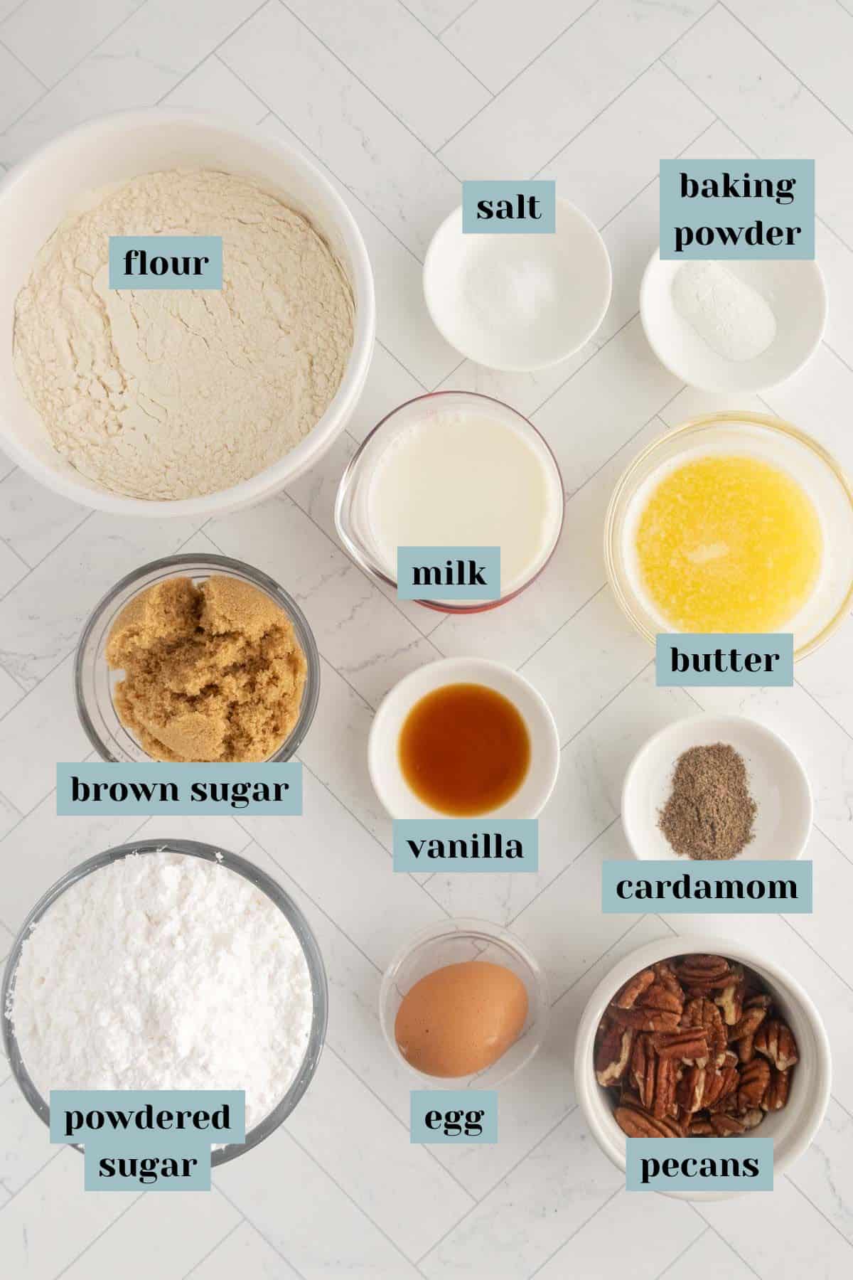 Top-down view of labeled baking ingredients in bowls, including flour, salt, baking powder, milk, butter, brown sugar, vanilla, cardamom, powdered sugar, egg, and pecans.