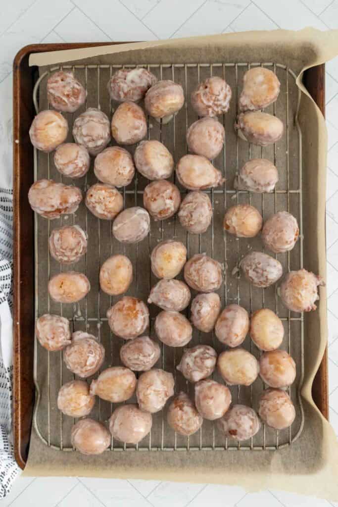 A baking tray lined with parchment paper holds a wire rack topped with dozens of round, glazed donut holes.