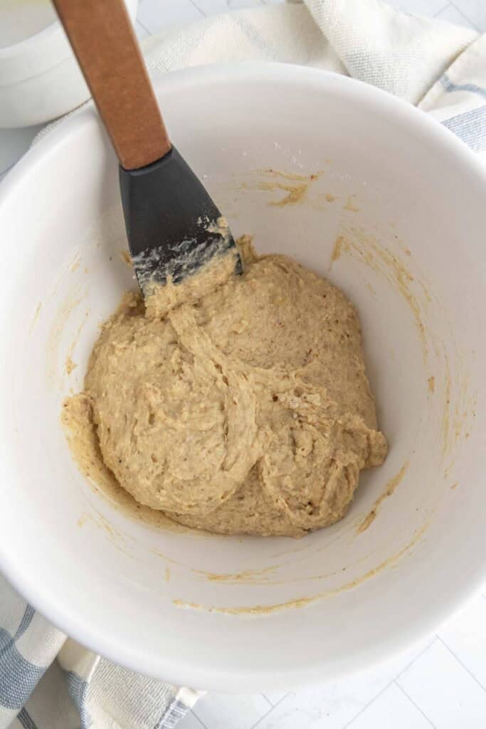 A mixing bowl containing thick, light brown dough being stirred with a black and wooden spatula, placed on a white and blue cloth.