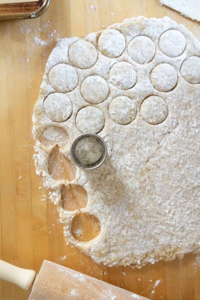 Round biscuit dough is being cut from a floured sheet with a metal cutter on a wooden surface. A rolling pin and dough scraps are visible nearby.