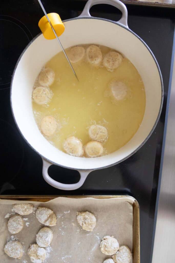 A pot of oil with dough balls frying and a thermometer attached; more dough balls rest on a parchment-lined tray nearby.