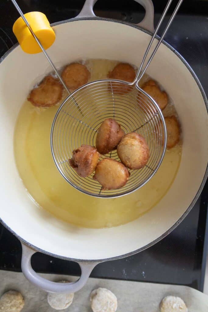A slotted spoon lifts several golden-brown dough balls from hot oil in a large pot; more dough balls fry in the oil below.