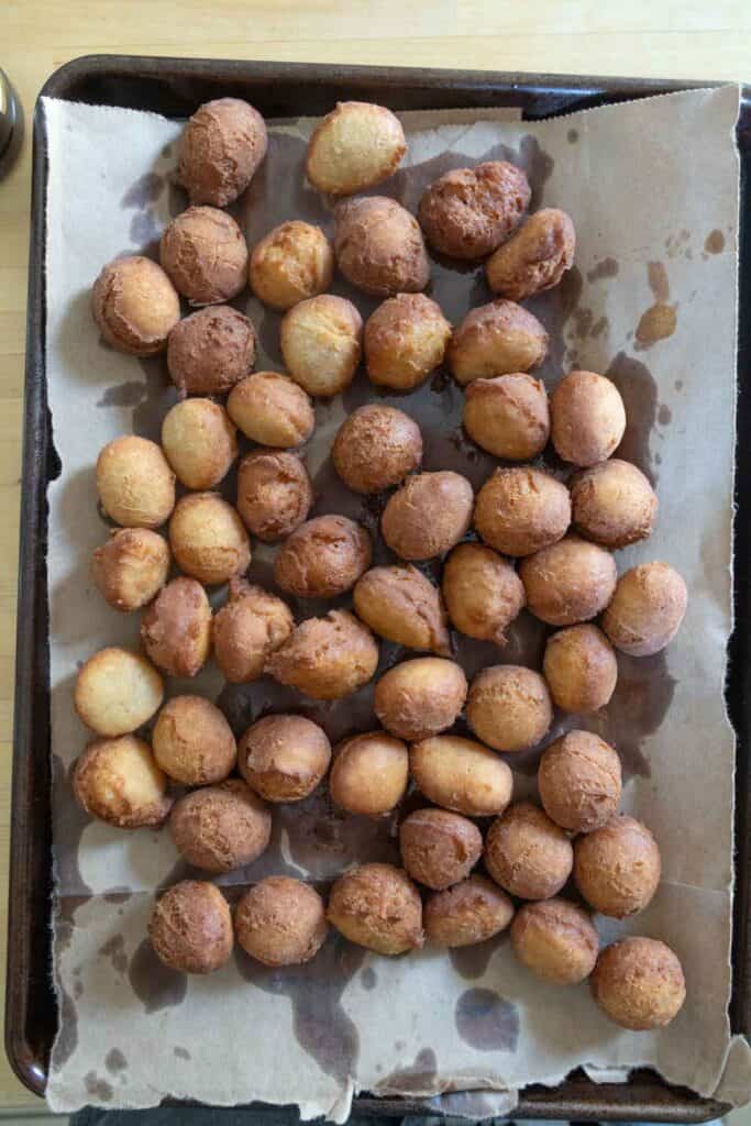 A baking sheet lined with parchment paper holds about 50 small, round, golden-brown fried dough balls with some oil spots visible on the paper.