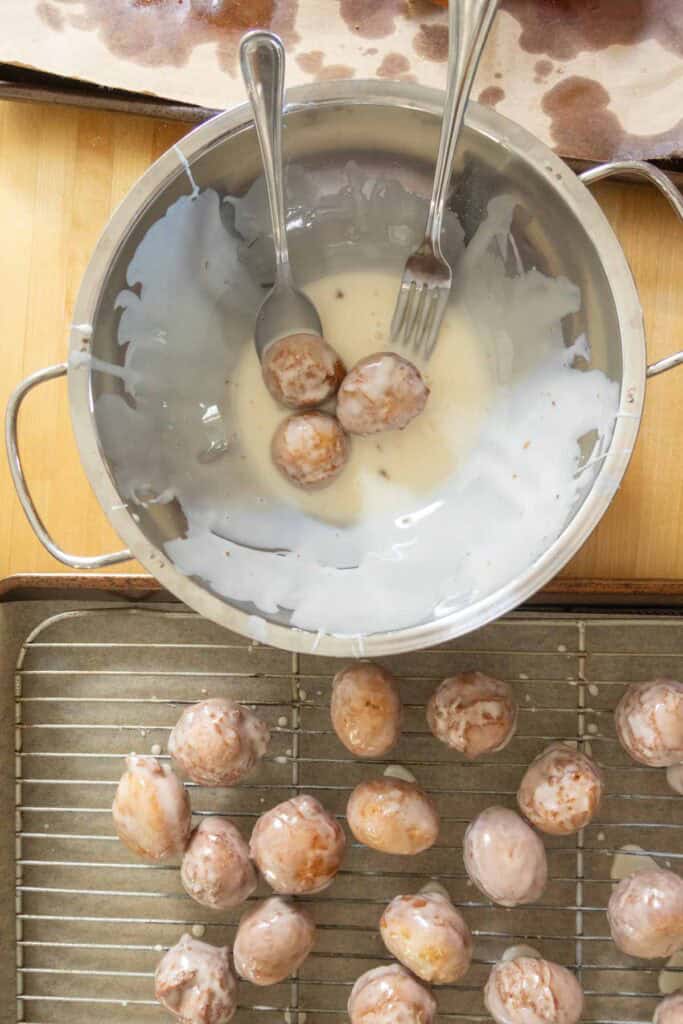 A metal bowl with glaze, two utensils, and doughnut holes being coated; some glazed doughnut holes are drying on a wire rack below.