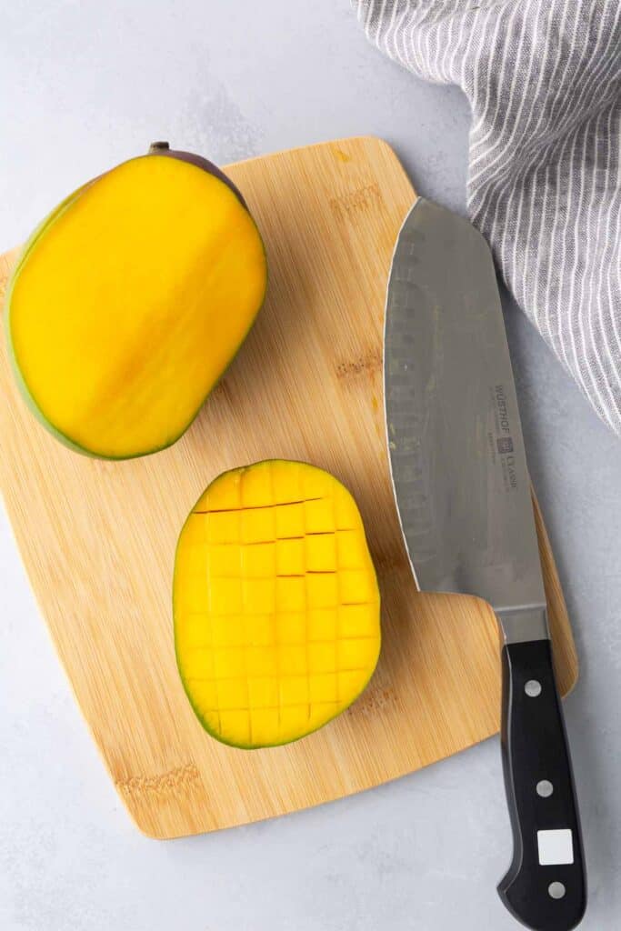 A halved mango on a wooden cutting board, one half diced in a grid pattern, with a large chef's knife and a striped towel nearby.