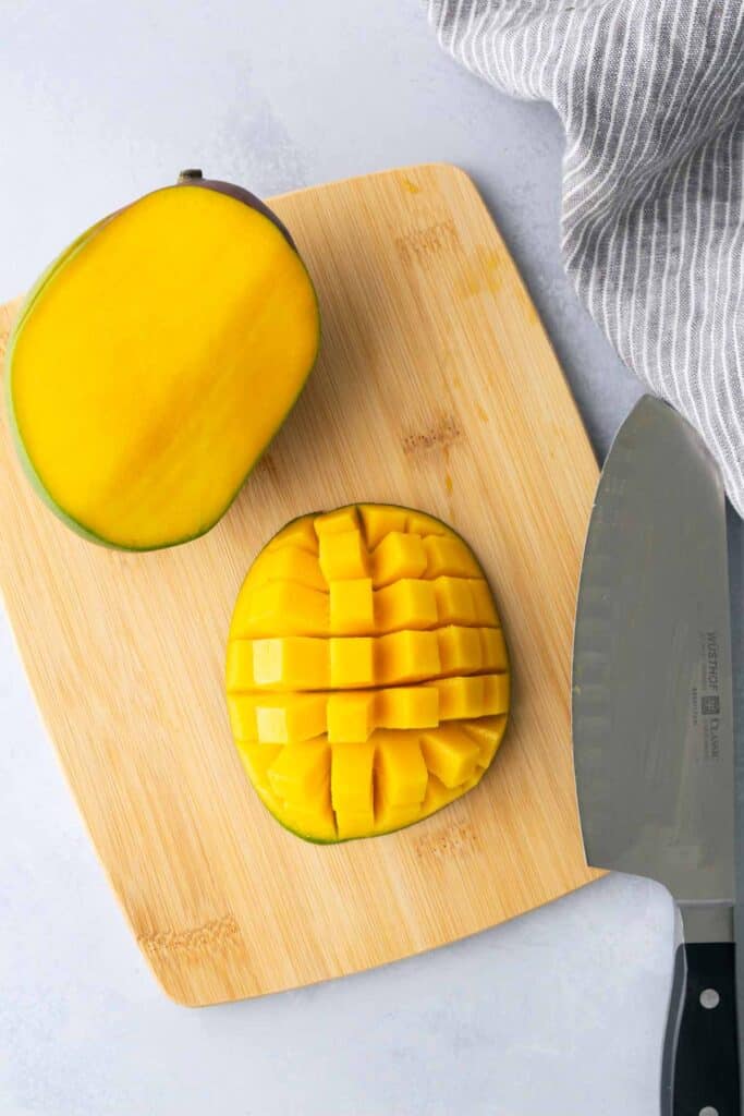 A mango half diced on a wooden cutting board, with the other half uncut; a knife and striped kitchen towel are next to the board.