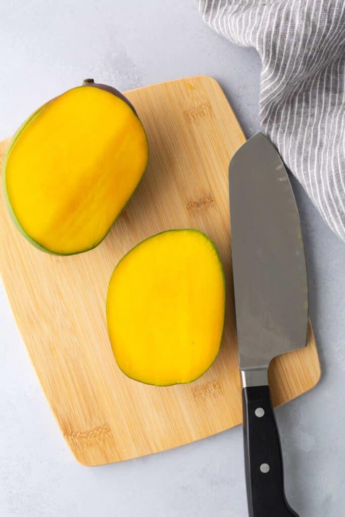A sliced mango with a chef’s knife on a wooden cutting board, next to a striped gray cloth.