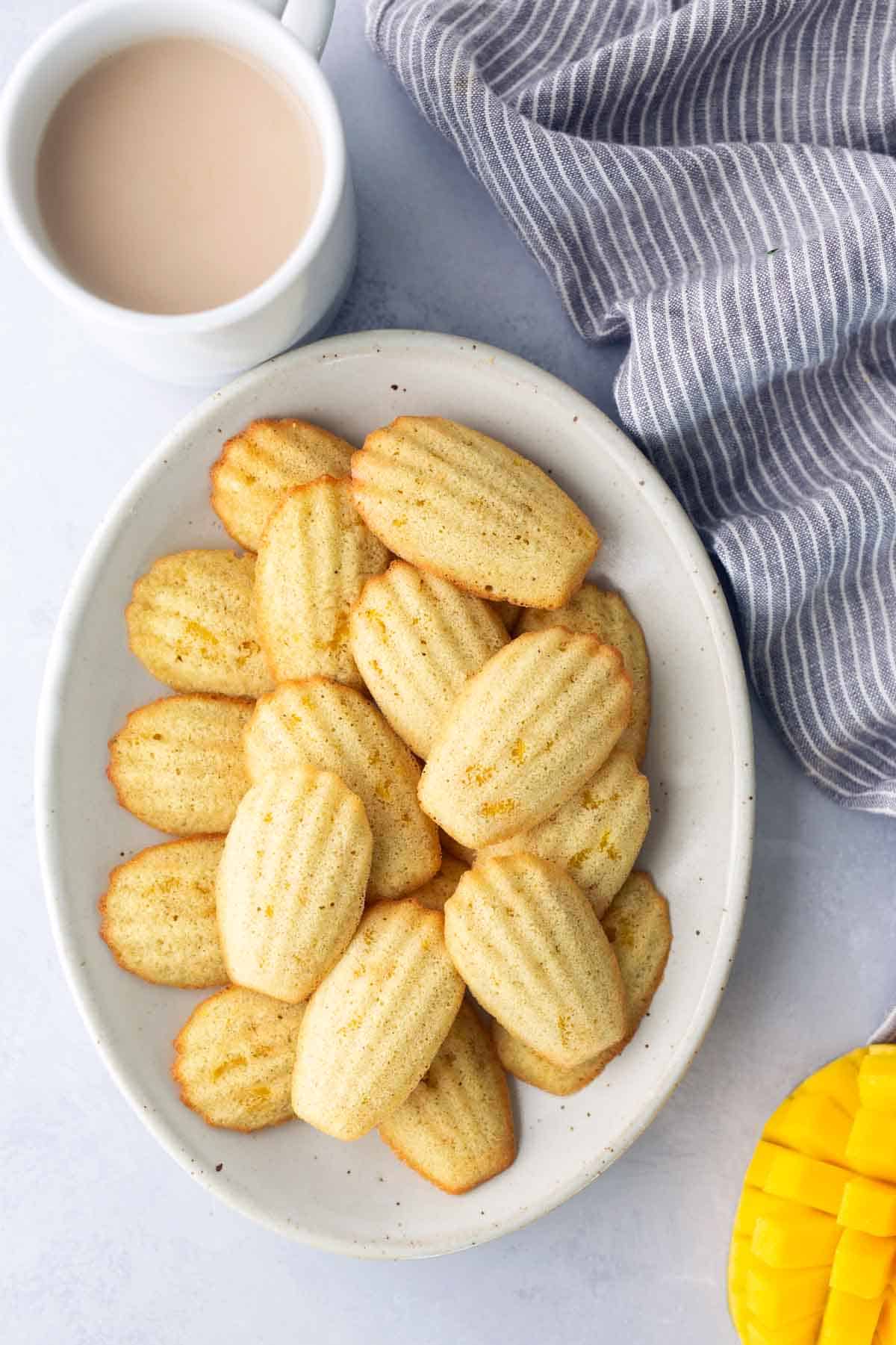 A white plate with madeleine cookies next to a cup of coffee or tea and a folded striped cloth on a light surface.