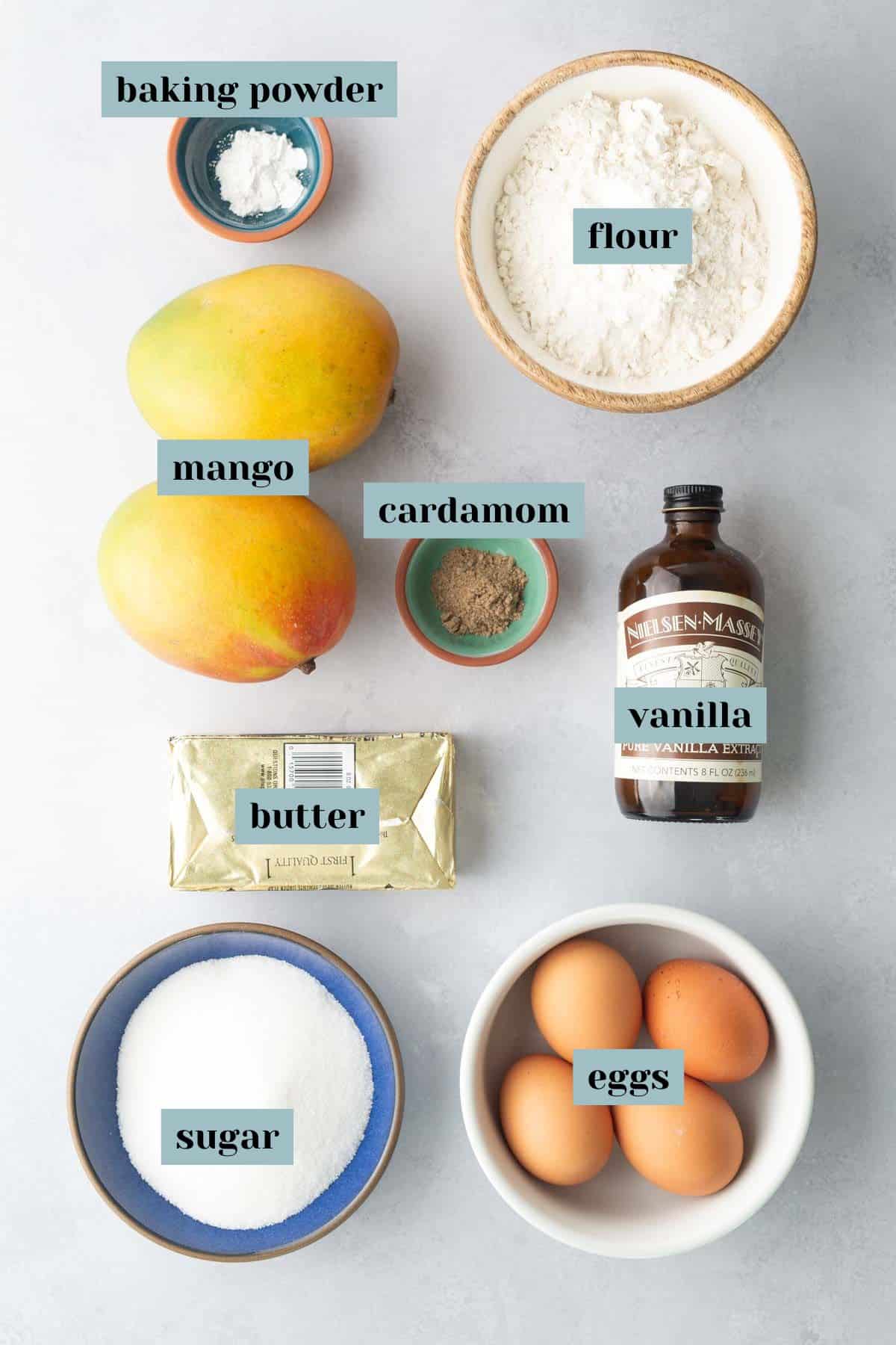 Top-down view of labeled baking ingredients: baking powder, flour, mango, cardamom, vanilla, butter, sugar, and eggs arranged on a light surface.