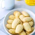 A white plate of madeleine cookies on a striped cloth, with a cup of coffee and two yellow mango-shaped objects in the background.