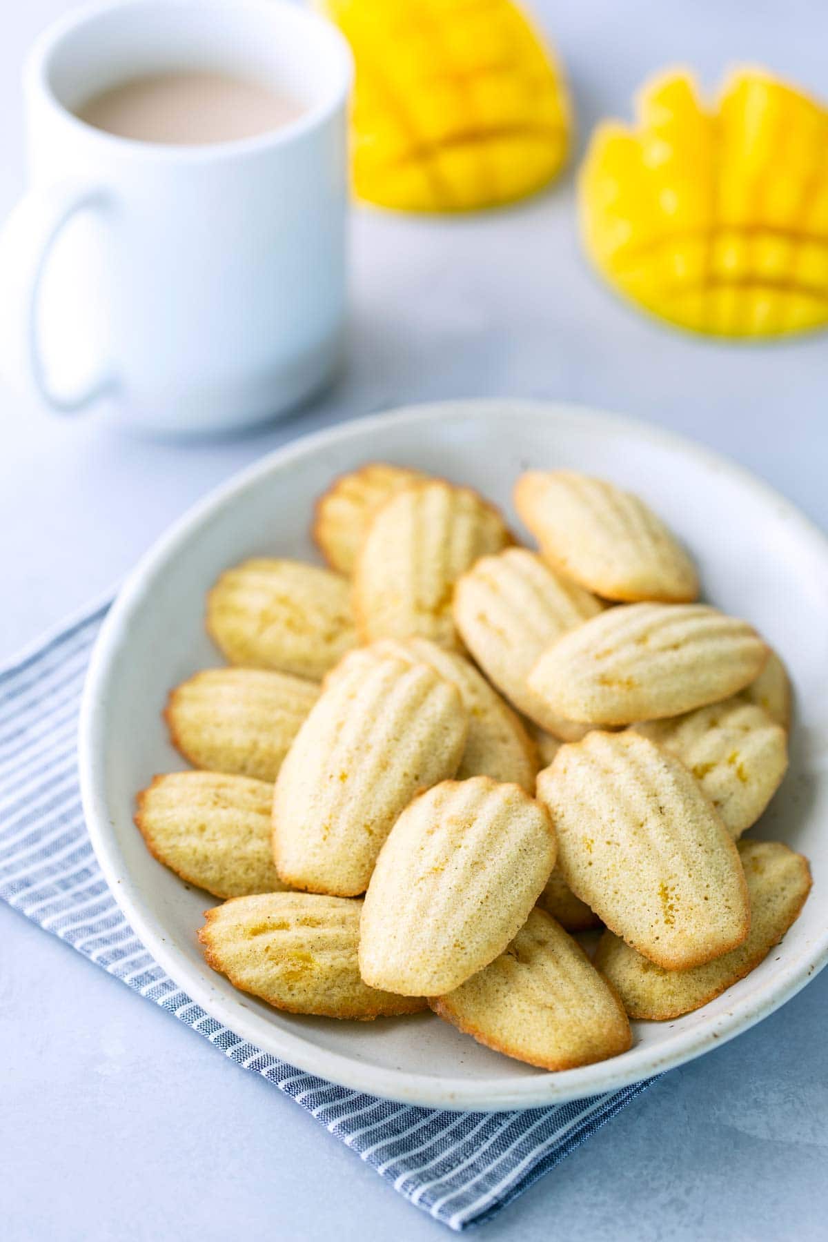 A white plate of madeleine cookies on a striped cloth, with a cup of coffee and two yellow mango-shaped objects in the background.
