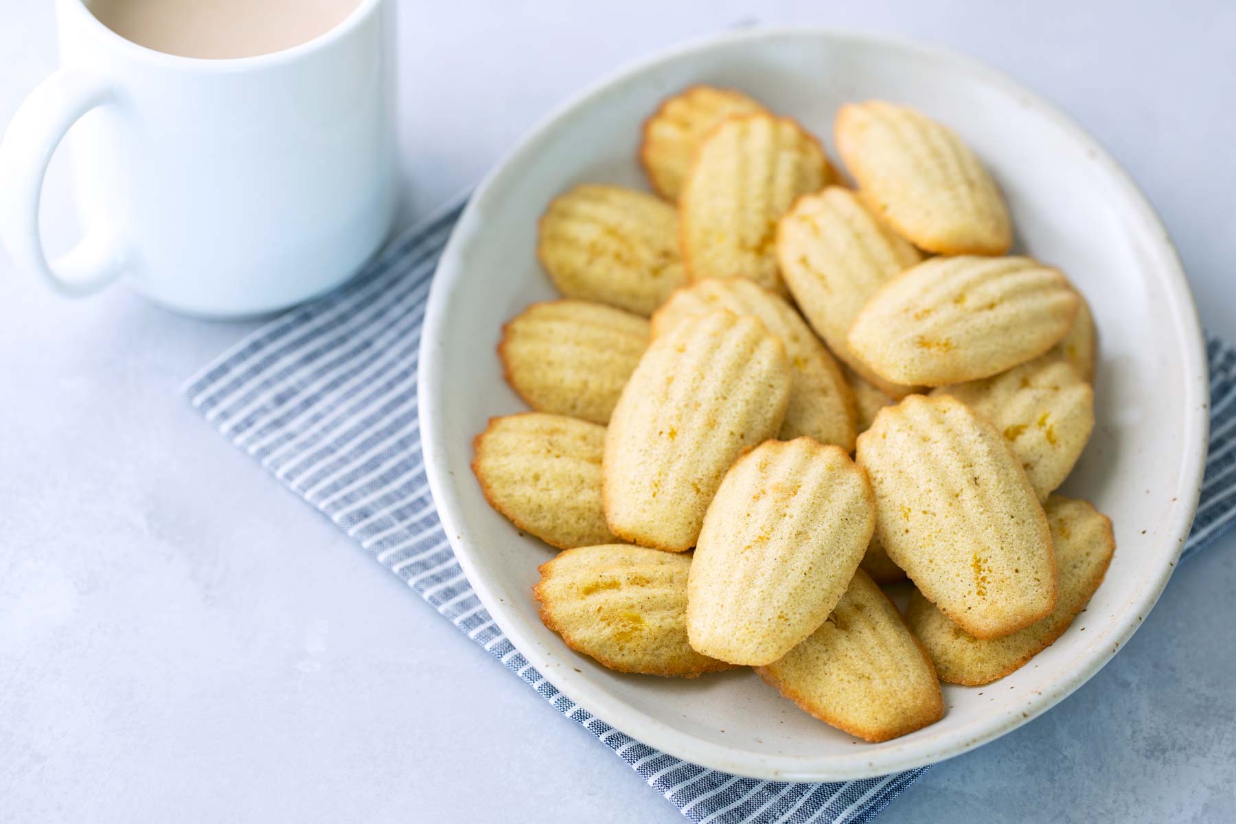 A plate of madeleine cookies on a striped cloth napkin next to a white mug filled with a light-colored beverage.