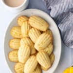 A plate of madeleine cookies sits next to a cup of coffee, a striped napkin, and sliced mango; text reads "Mango Madeleines.