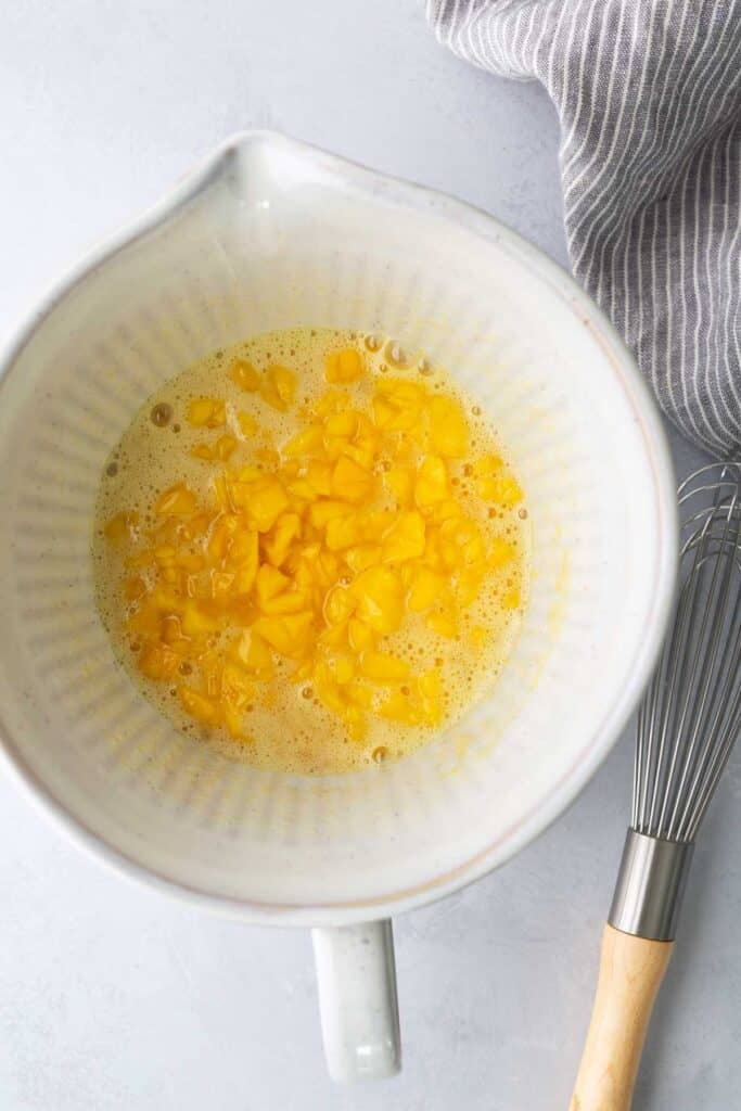 A white mixing bowl with beaten eggs and chopped yellow bell peppers, next to a whisk and a gray striped cloth on a light surface.