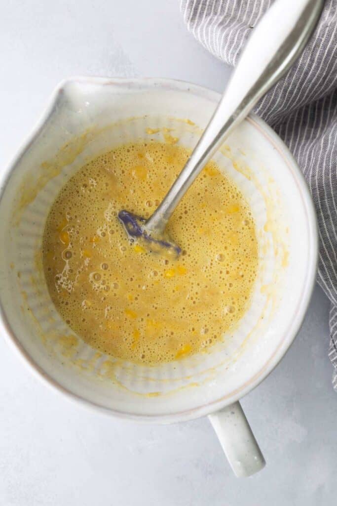 A white mixing bowl with a metal spoon and a mixture of beaten eggs; a gray striped cloth is partially visible on the side.