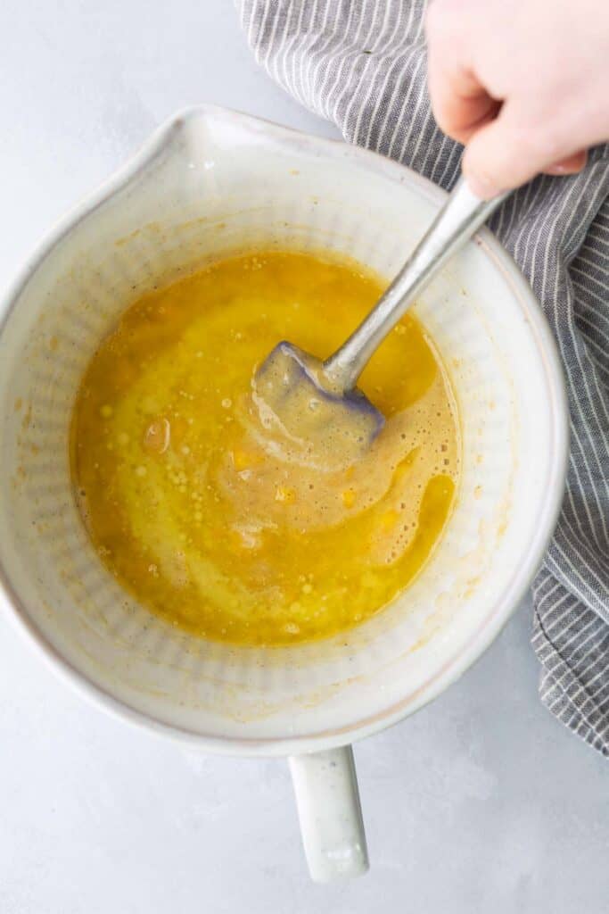 Hand stirring a yellow liquid mixture with a spatula in a white mixing bowl next to a gray striped cloth on a light surface.