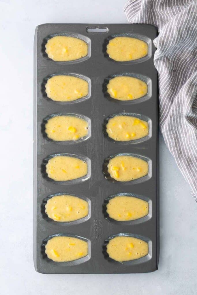 A madeleine pan filled with unbaked batter, some with visible chunks, next to a striped dish towel on a light surface.