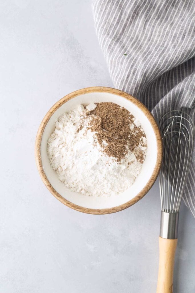 A wooden bowl with flour and ground pepper next to a metal whisk and a gray striped cloth on a light surface.