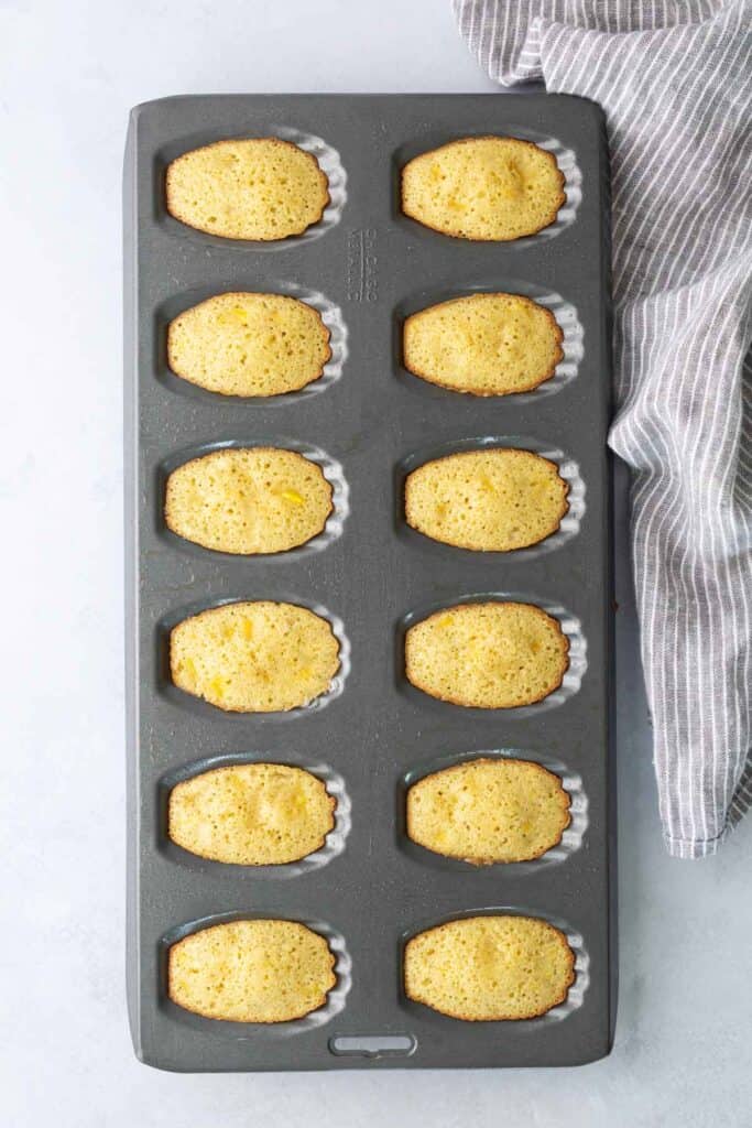 A madeleine pan filled with twelve freshly baked madeleine cakes, resting next to a gray and white striped kitchen towel.