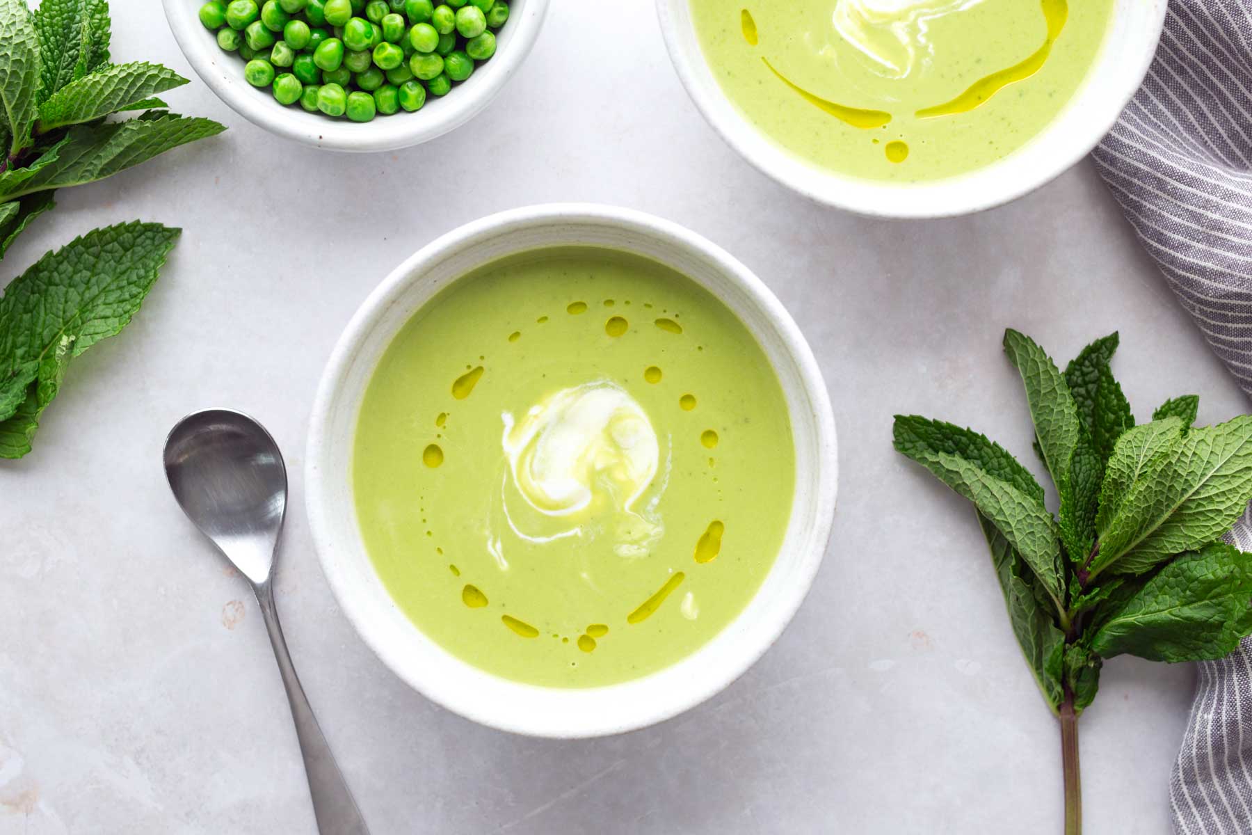 A bowl of green pea soup with a swirl of cream and a drizzle of oil, next to a spoon, fresh mint leaves, and a bowl of peas on a light surface.