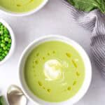 Two bowls of green pea soup topped with a swirl of cream and a drizzle of oil, surrounded by a spoon, fresh mint leaves, and a small bowl of peas.