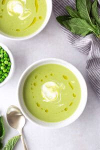 Two bowls of green pea soup topped with a swirl of cream and a drizzle of oil, surrounded by a spoon, fresh mint leaves, and a small bowl of peas.