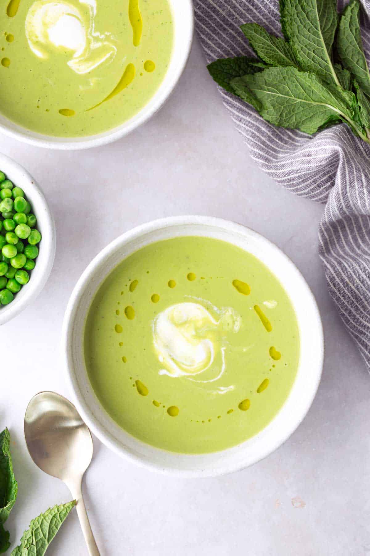 Two bowls of green pea soup topped with a swirl of cream and a drizzle of oil, surrounded by a spoon, fresh mint leaves, and a small bowl of peas.