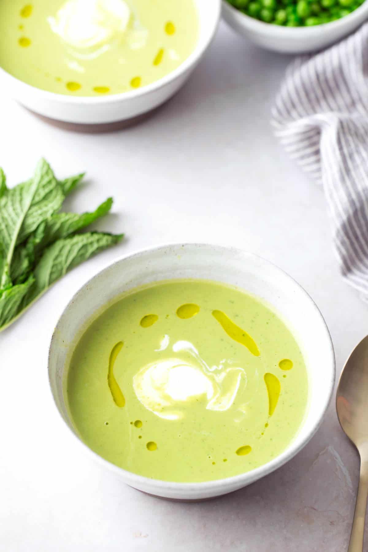 A bowl of green soup with a swirl of cream and a drizzle of oil on top, garnished with mint leaves, next to a spoon and a bowl of green peas.