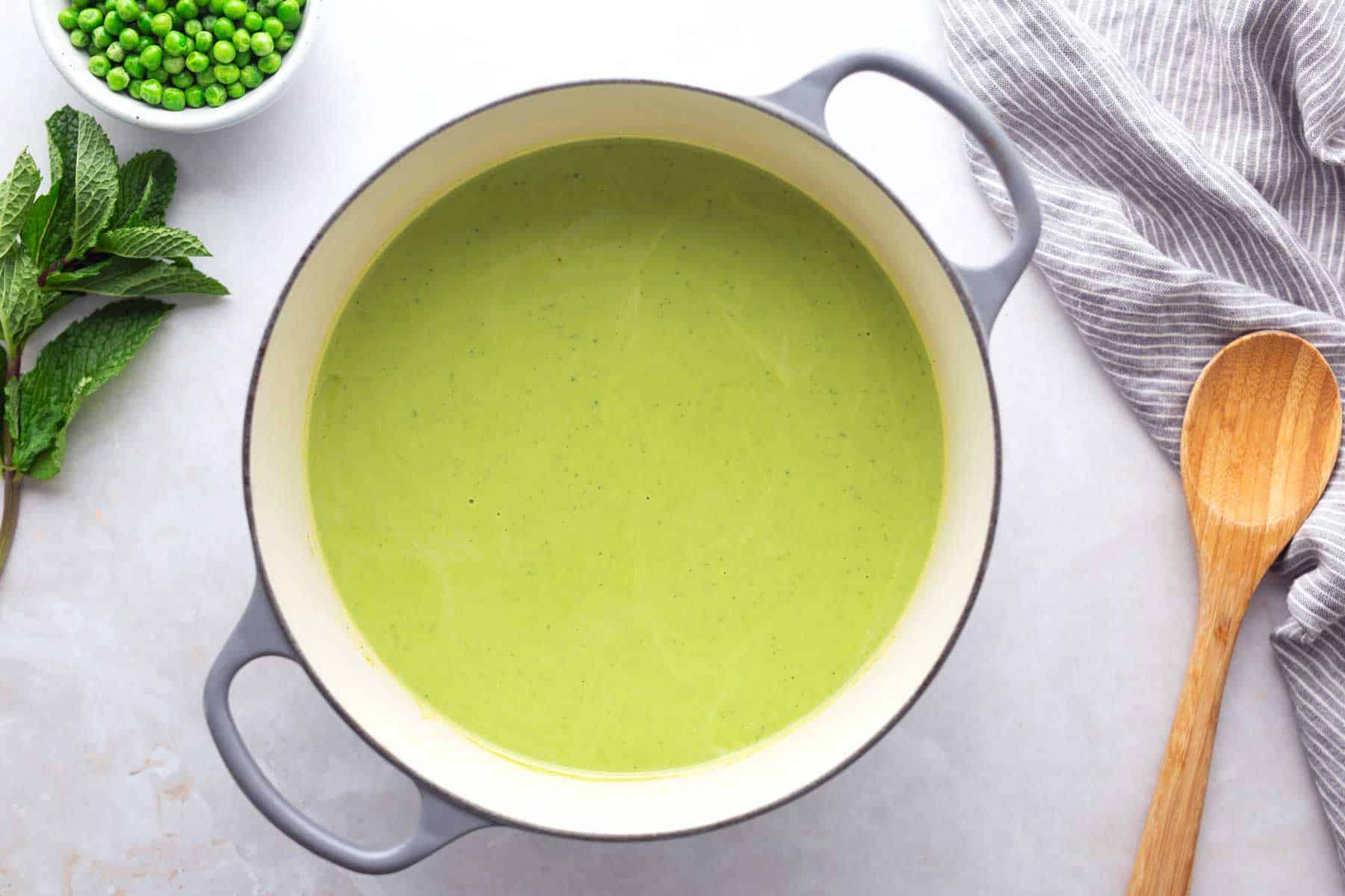 A pot of green soup sits on a countertop next to a wooden spoon, a bowl of peas, fresh mint, and a striped kitchen towel.