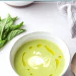 A bowl of mint pea soup topped with a swirl of cream and a drizzle of oil, garnished with fresh mint leaves beside the bowl.