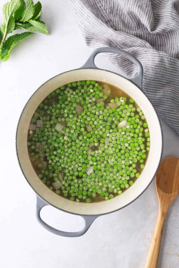 A pot filled with green peas, diced onions, and broth sits on a light surface next to a wooden spoon, striped towel, and fresh mint leaves.