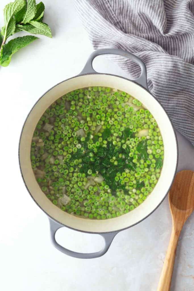 A pot of soup with chopped green beans and herbs on a light surface, next to a wooden spoon, striped towel, and fresh mint leaves.