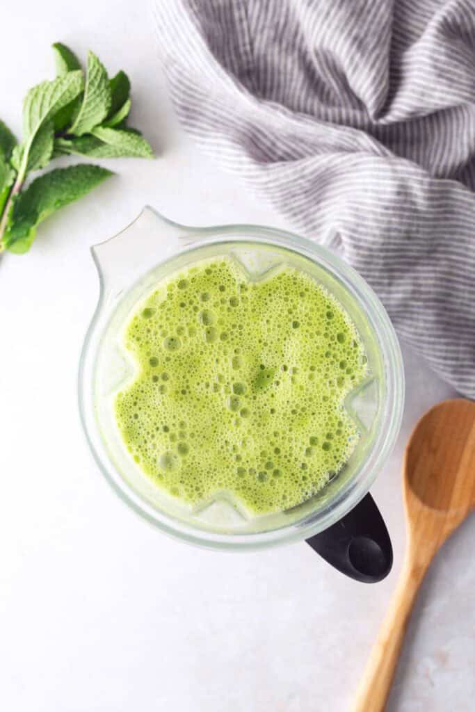 A top view of a blender filled with green smoothie, next to a wooden spoon, fresh mint leaves, and a gray-striped cloth on a light surface.