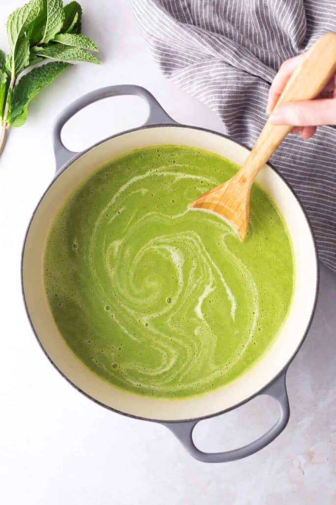 A hand stirs a pot of creamy green soup with a wooden spoon. Fresh mint leaves and a striped cloth are nearby on a light surface.