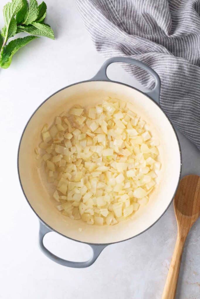 Diced onions are sautéing in a white pot on a countertop, with a wooden spoon, a striped cloth, and fresh mint leaves nearby.