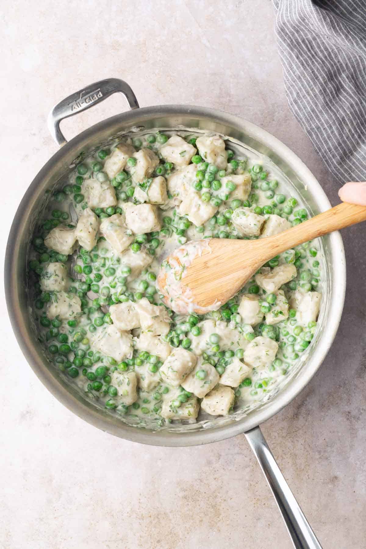 A pan filled with creamy chicken and peas being stirred with a wooden spoon.