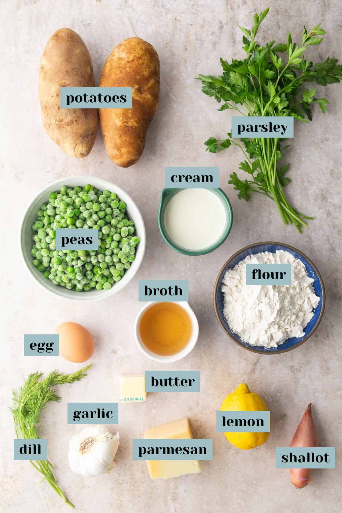 Overhead view of labeled ingredients for a recipe, including potatoes, parsley, peas, cream, flour, broth, egg, butter, garlic, parmesan, dill, lemon, and shallot on a light surface.