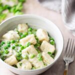 A bowl of gnocchi with green peas in a creamy white sauce, placed on a light surface next to a fork and a striped cloth.