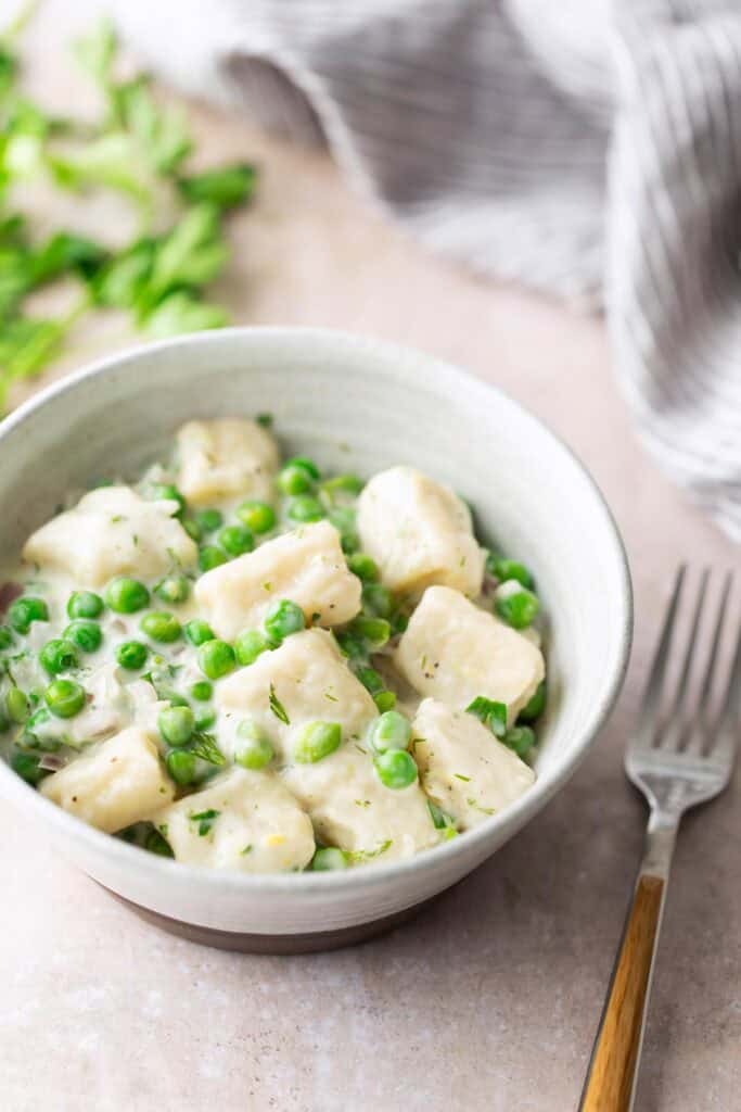 A bowl of gnocchi with green peas in a creamy white sauce, placed on a light surface next to a fork and a striped cloth.