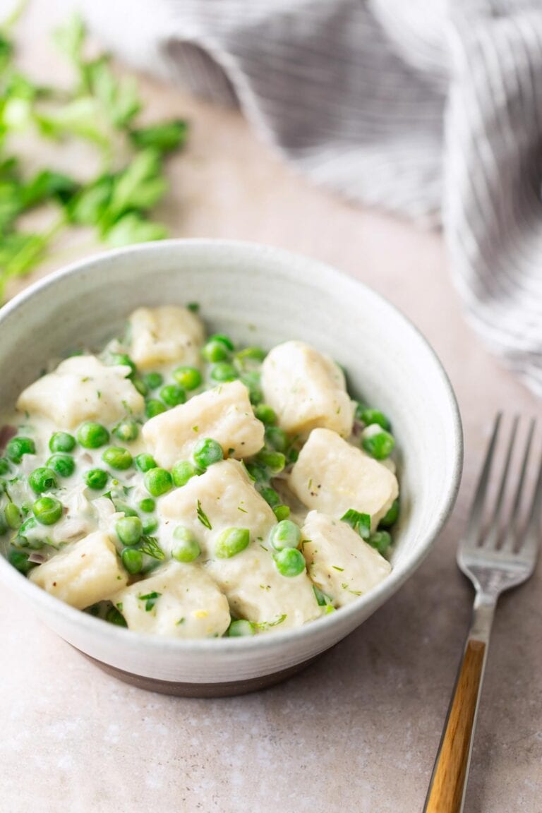 A bowl of gnocchi with green peas in a creamy white sauce, placed on a light surface next to a fork and a striped cloth.