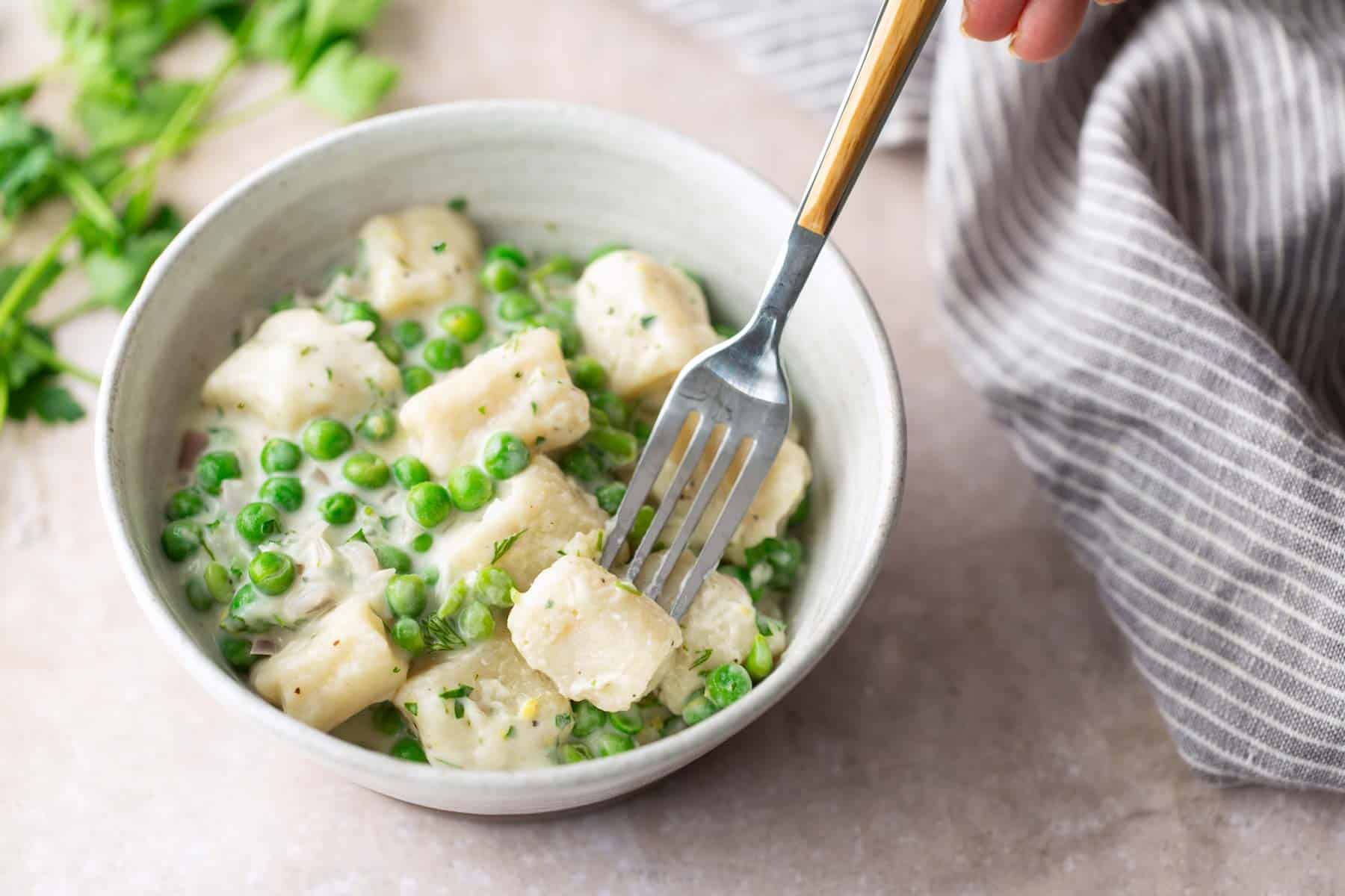 A hand holds a fork over a bowl of gnocchi with green peas in a creamy sauce, next to a striped cloth and fresh herbs on a light surface.
